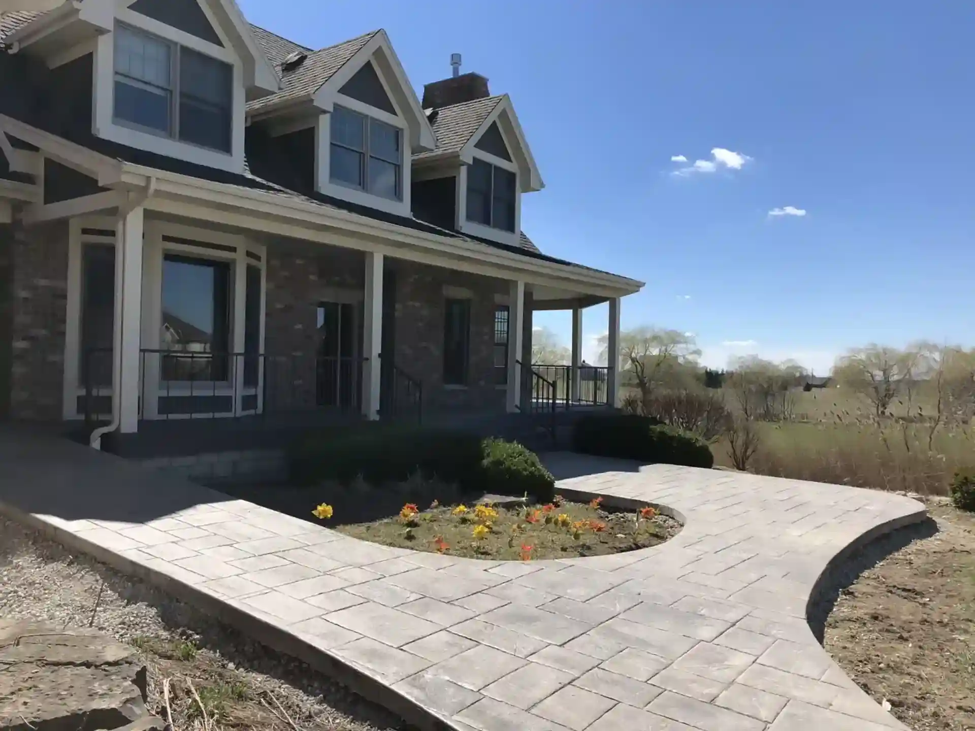 custom stone pathway leading to the entrance of a large home with a landscaped flower bed, showcasing a curved design and lush greenery in the surrounding area.