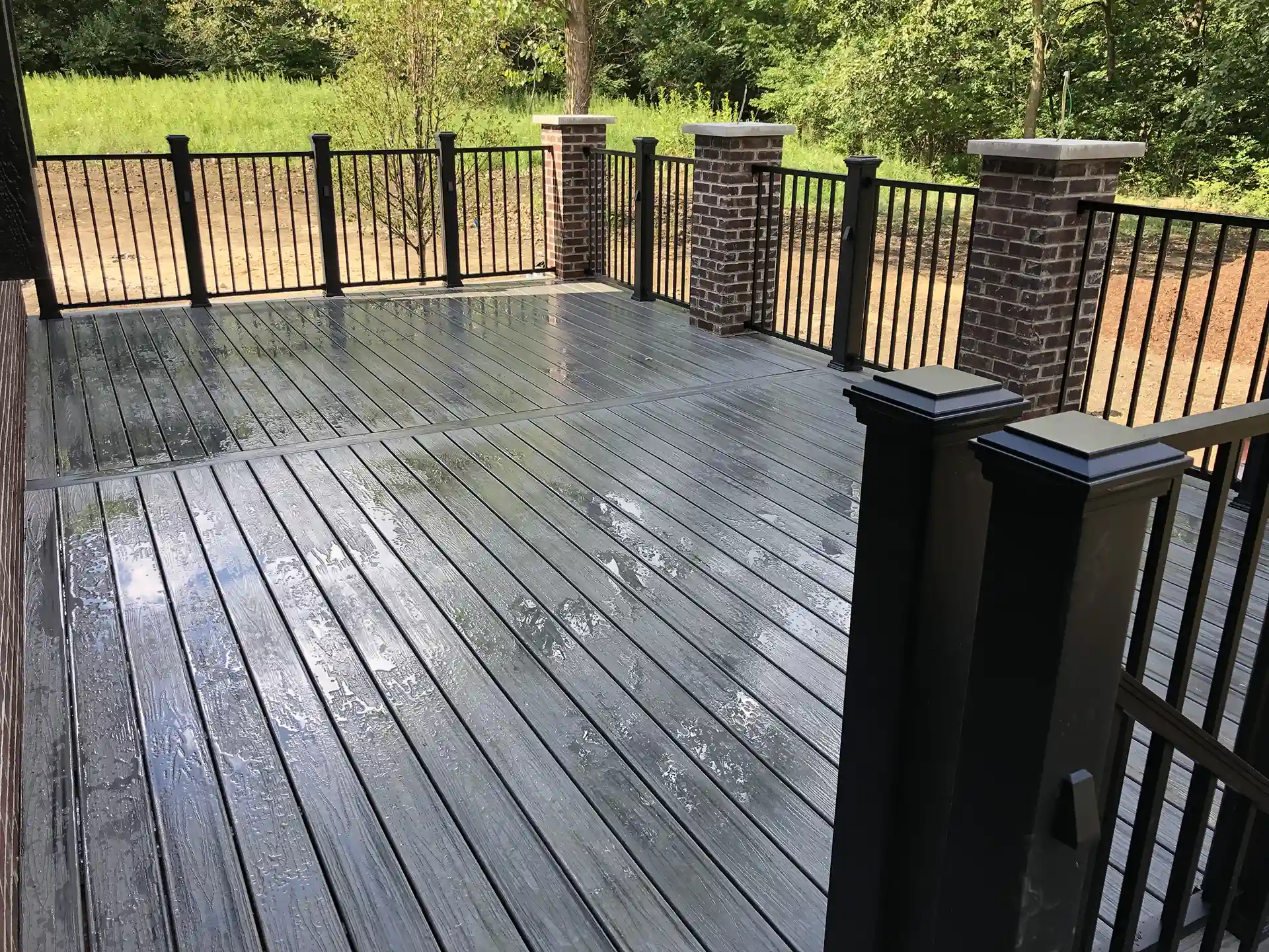 deck with composite flooring and black railings, bordered by brick columns and surrounded by greenery.
