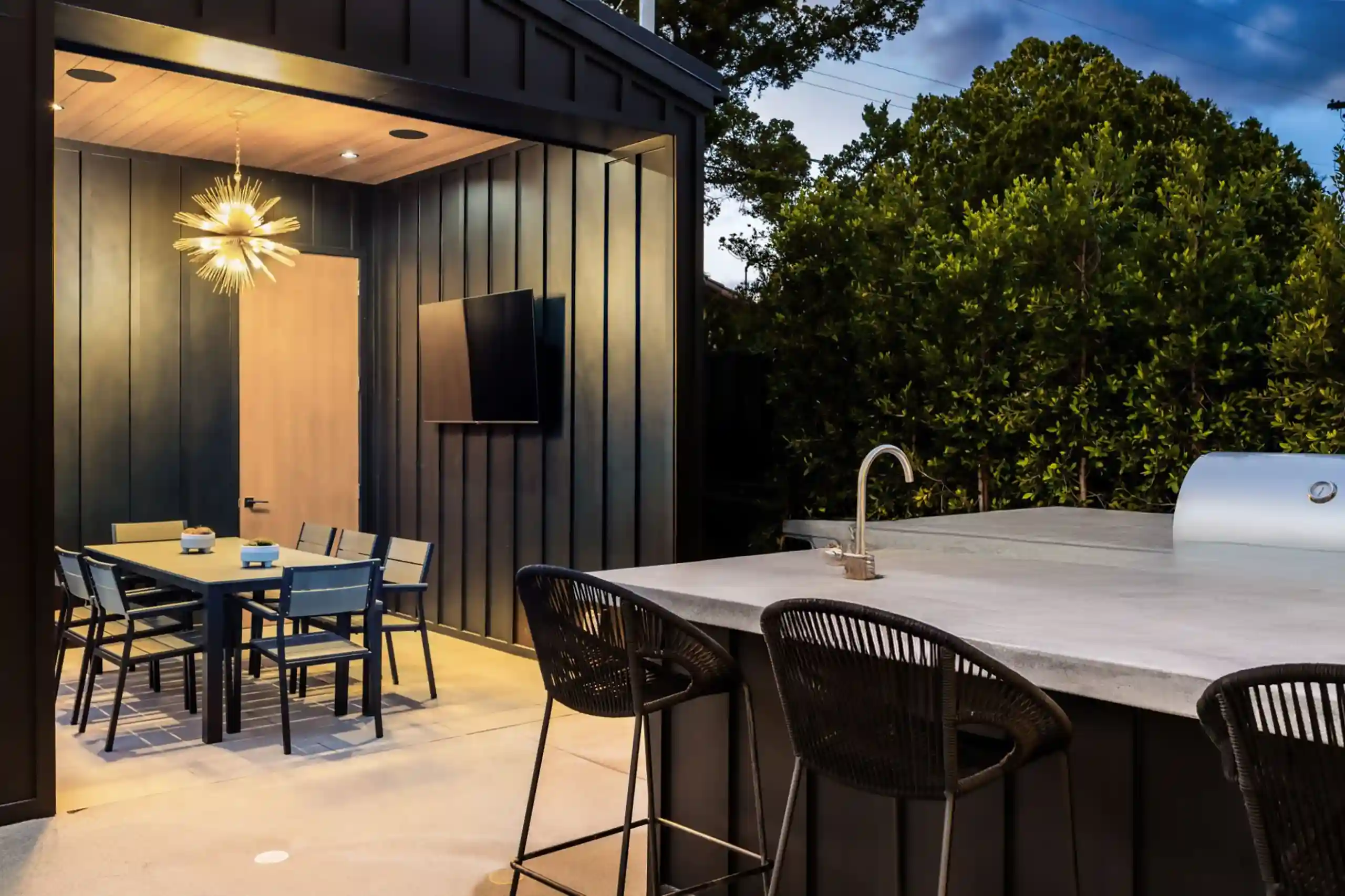 outdoor kitchen with built-in grill, sink, and bar seating next to a covered dining area with a table, chairs, ceiling light fixture, and wall-mounted television.