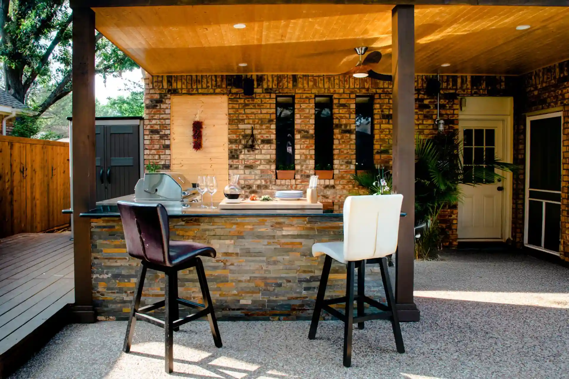 covered outdoor kitchen with built-in grill, stone bar island, two barstools, ceiling fan, and brick wall backdrop in a backyard patio setting.
