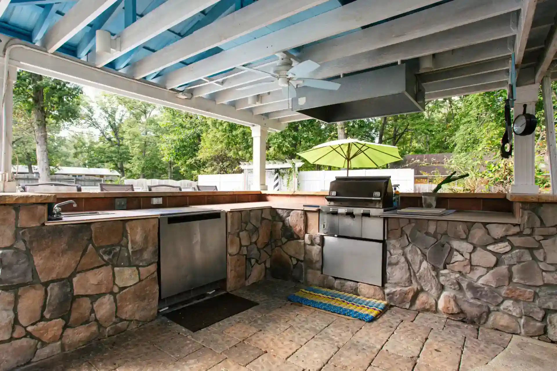 covered outdoor kitchen with stone countertops, built-in stainless steel appliances, ceiling fan, and a grill under an open rafter roof in a backyard setting.
