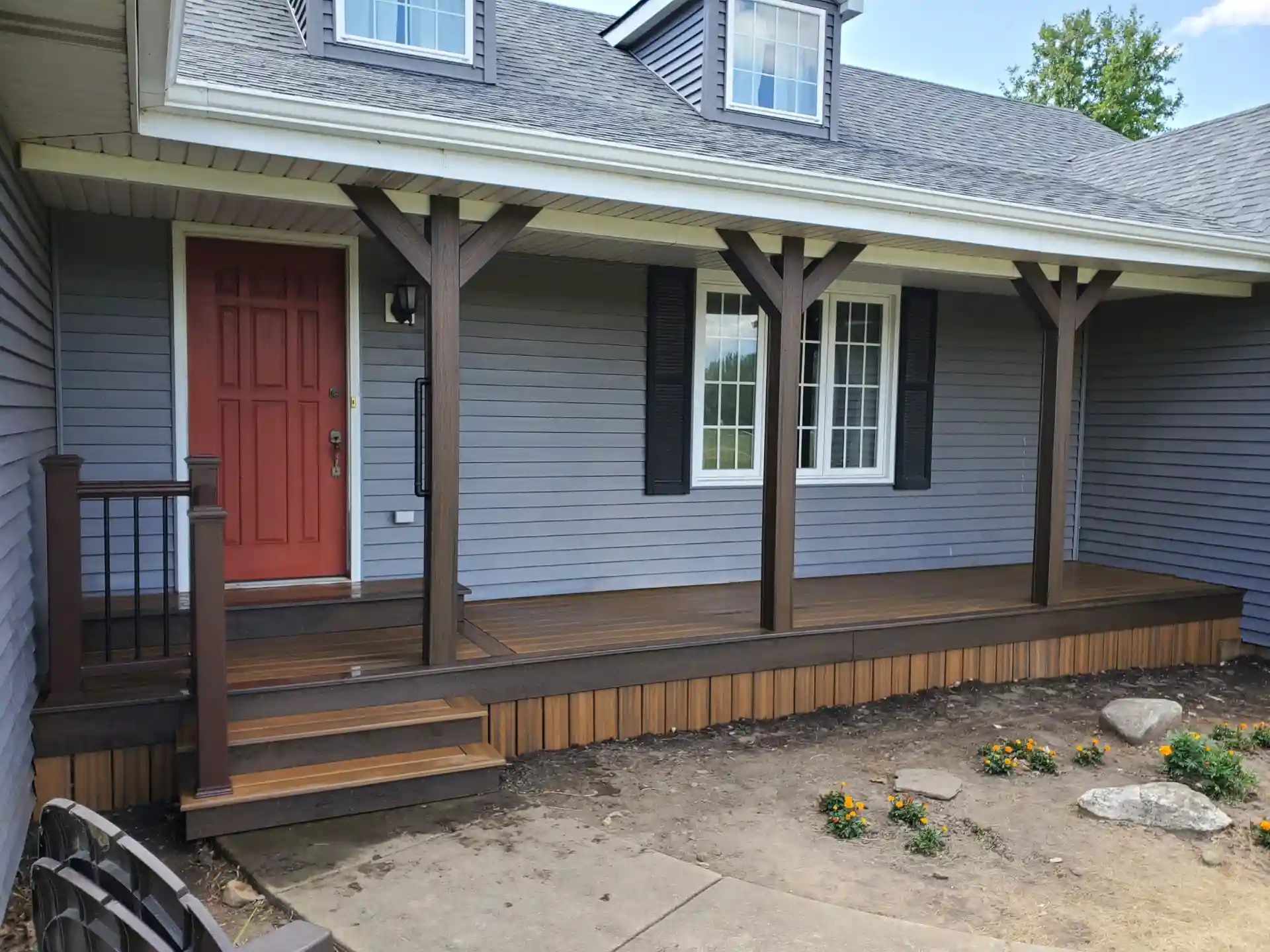 front porch with wood decking, wooden support columns, and a red front door. the porch is surrounded by landscaping with flowers and a gravel path.

