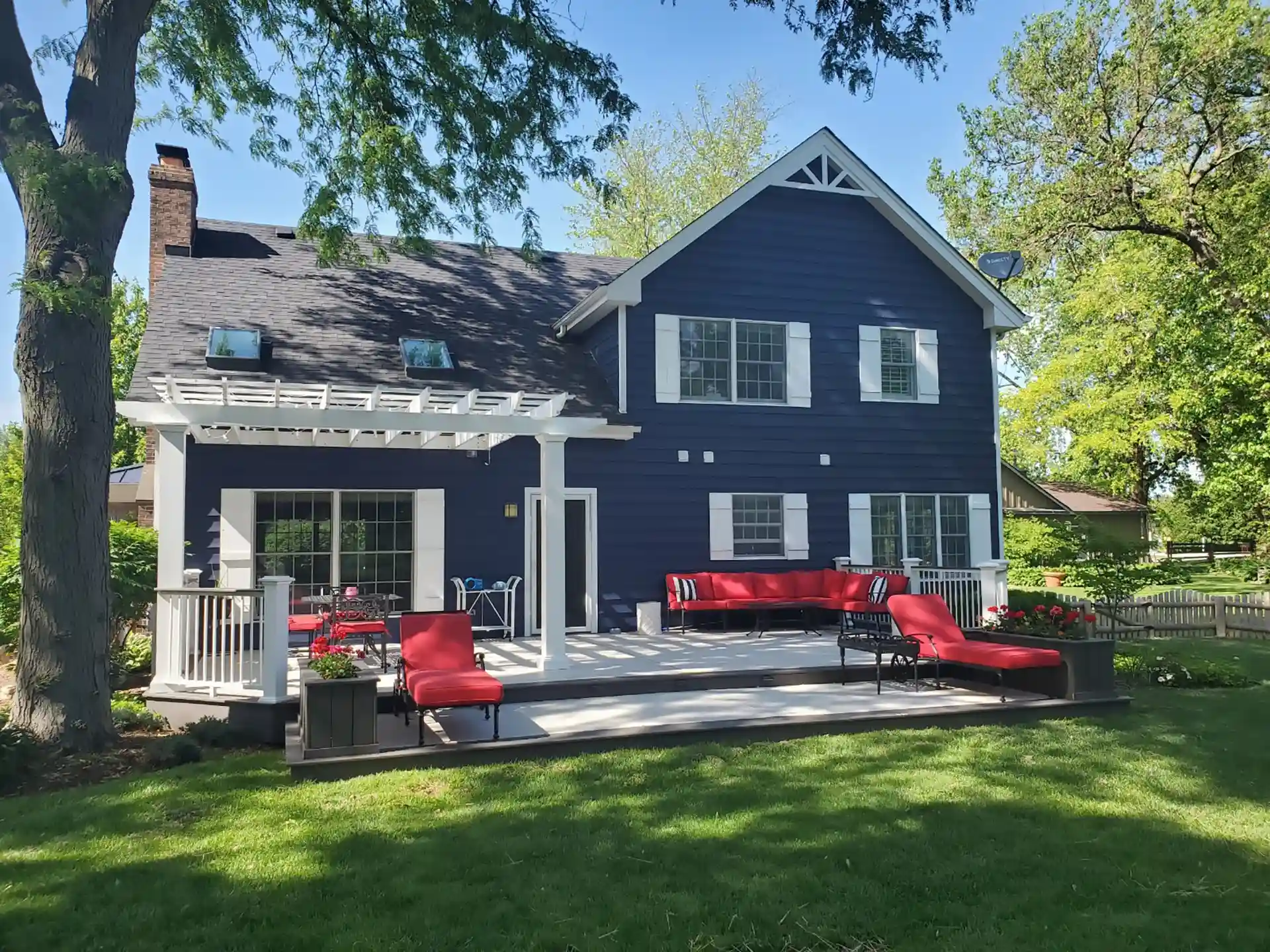 back porch with a large deck, red outdoor furniture, and a white pergola. the house has blue siding and white trim, with large windows and a well-maintained lawn.
