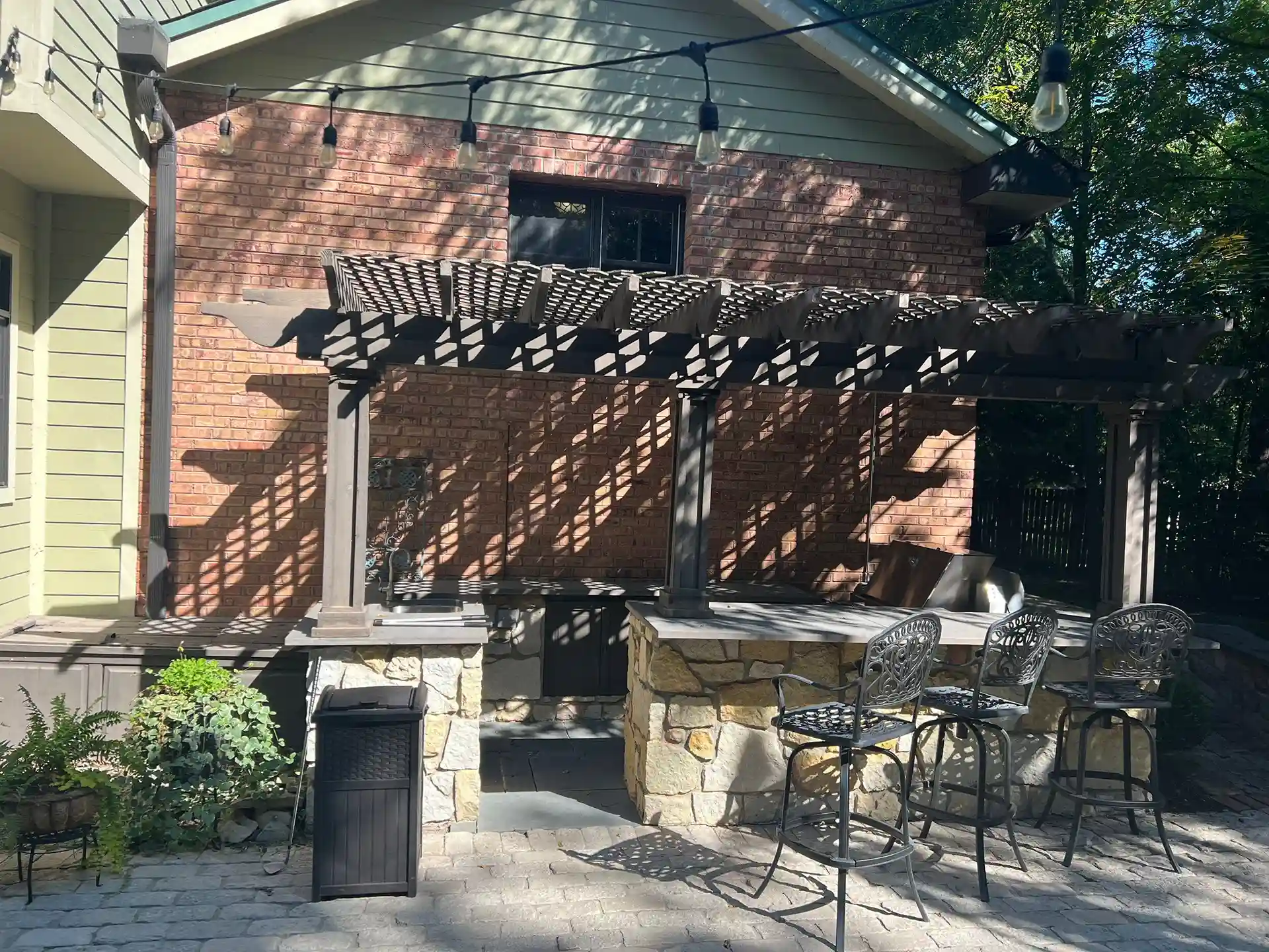 outdoor patio with a wooden pergola, stone bar counter, and metal barstools, set against a brick house with string lights.
