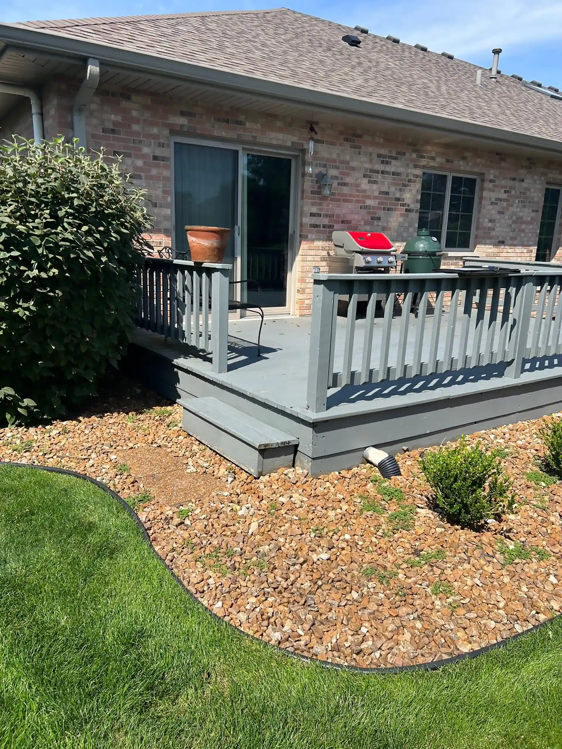 small gray wooden deck with a railing, outdoor grill, and potted plant attached to a brick house with a landscaped yard.
