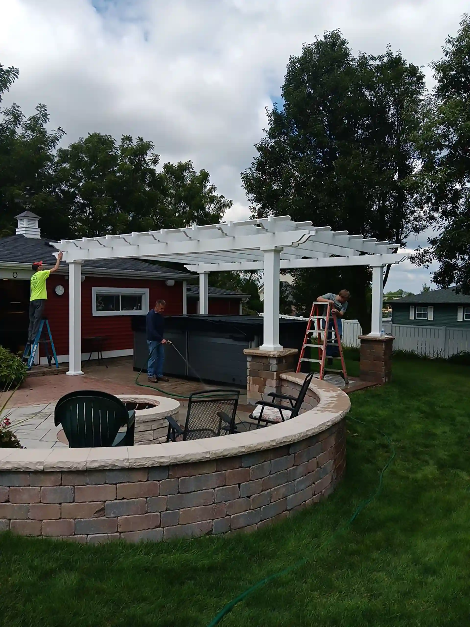 white pergola installation over a patio with a fire pit, surrounded by a brick wall, with workers assembling the structure.
